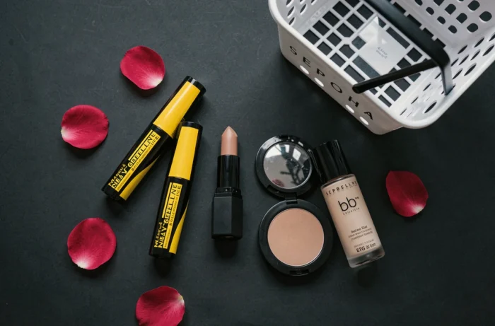 Plat posé de produits Maybelline best-sellers incluant mascara, rouge à lèvres et fond de teint aux côtés d'un panier Sephora sur fond noir mat